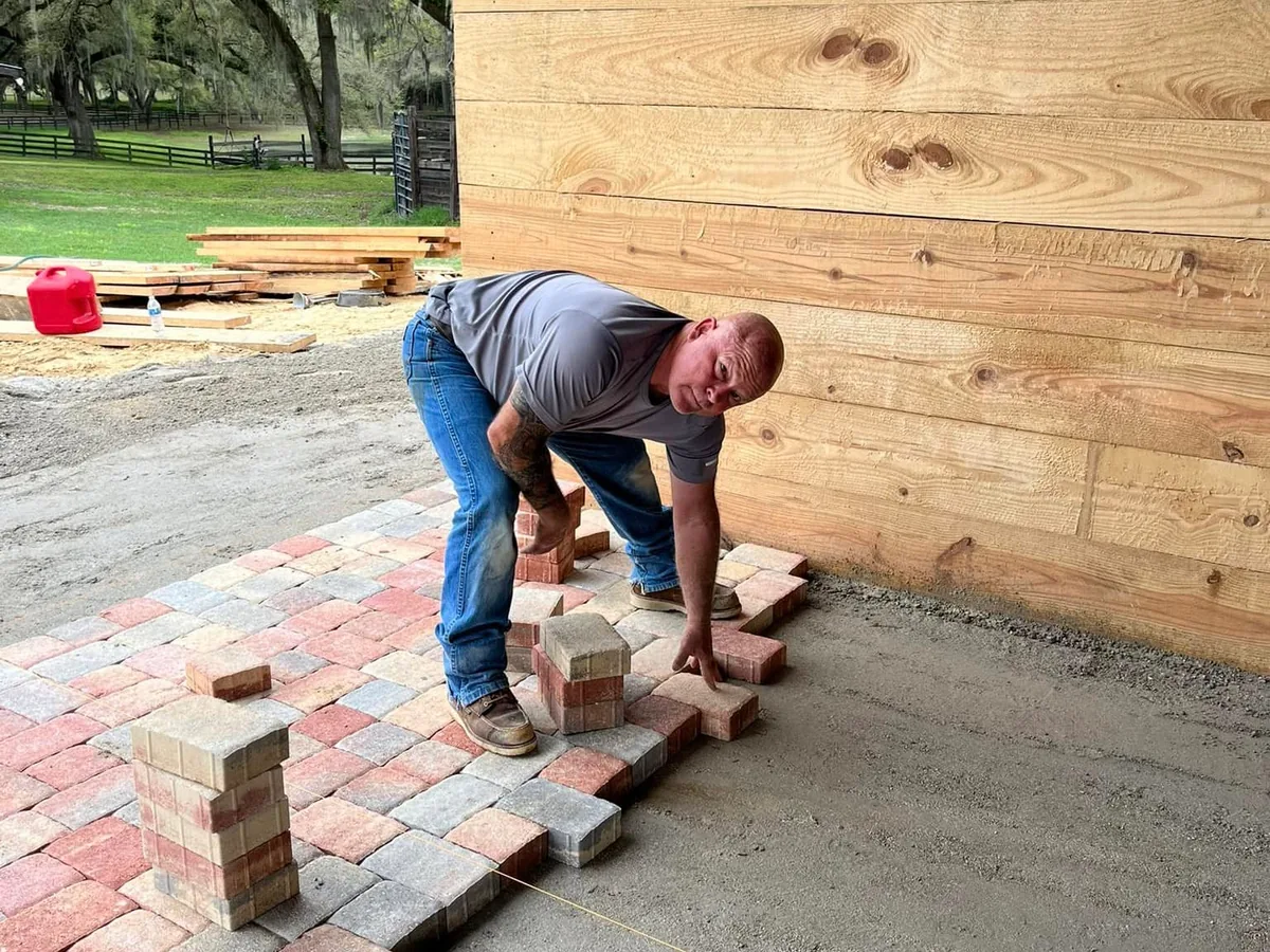 Crew laying tumbled pavers on a screeded base beside a barn