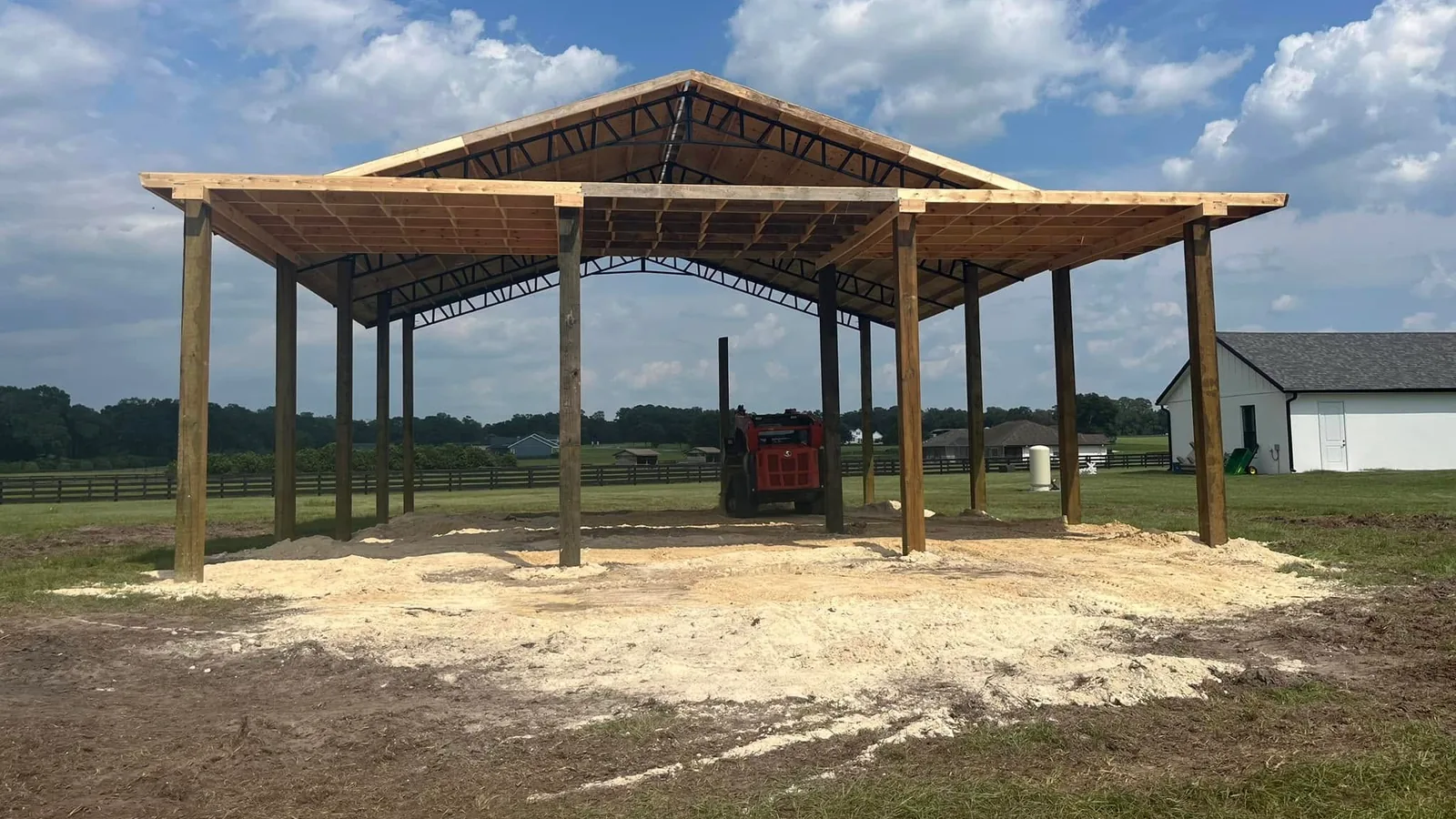 Pole barn pavilion with steel trusses on a sand pad beside board fenced pastures