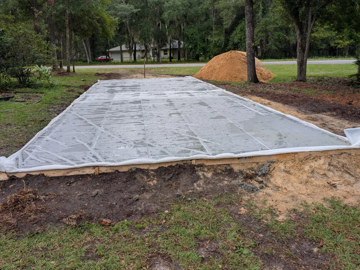 Freshly poured concrete pad curing under plastic with the forms still in place on a wooded lot