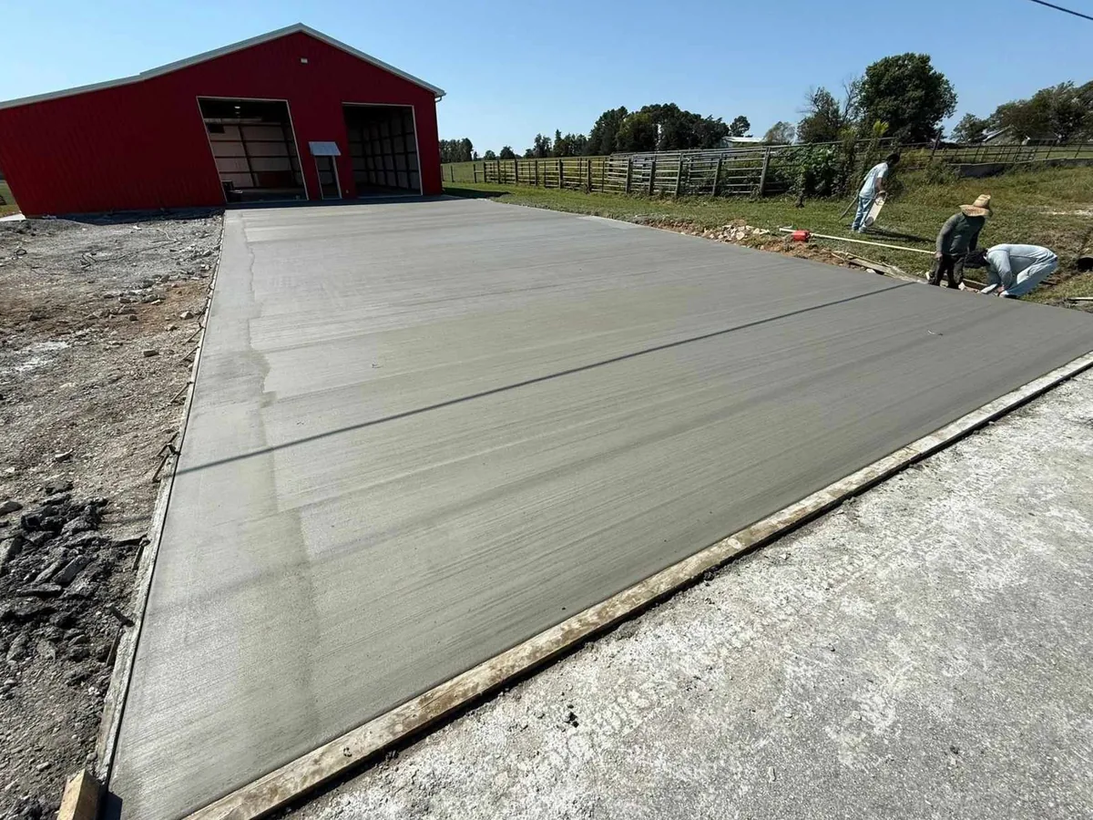 Crew finishing the edge of a wide new broom finish concrete driveway leading to a red metal barn