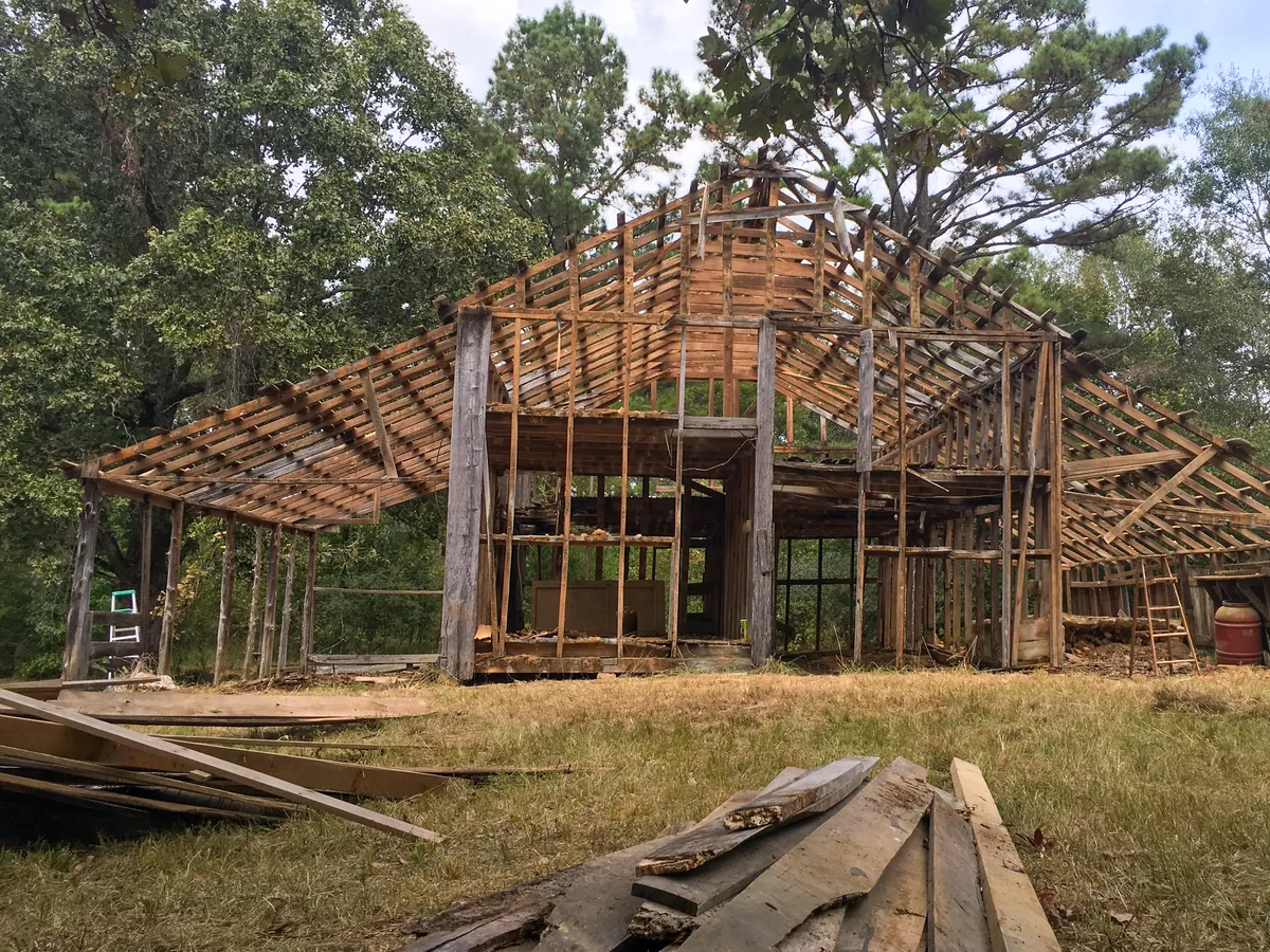 Old wooden barn partly torn down with its roof framing exposed and salvaged boards stacked in front
