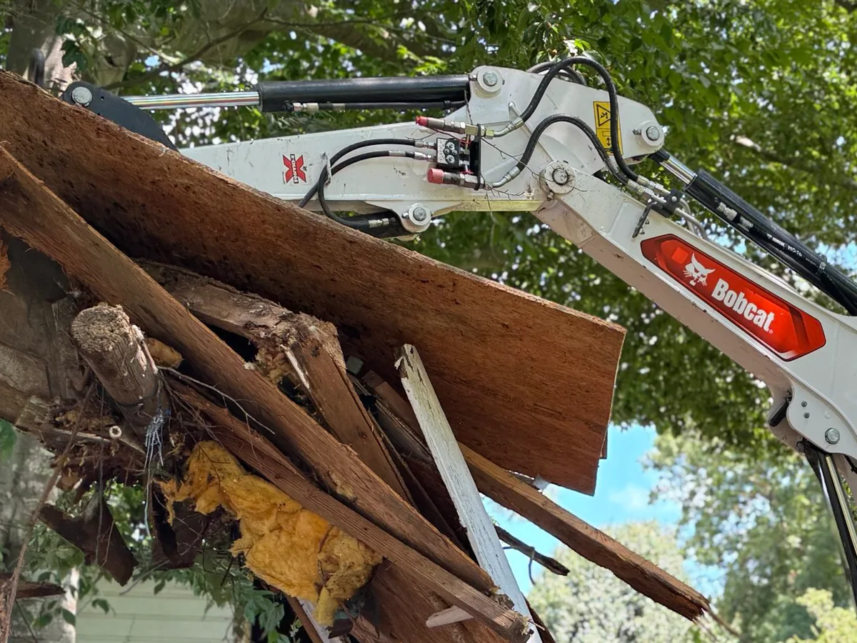 Excavator arm pulling apart the roof framing and insulation of an old wooden building