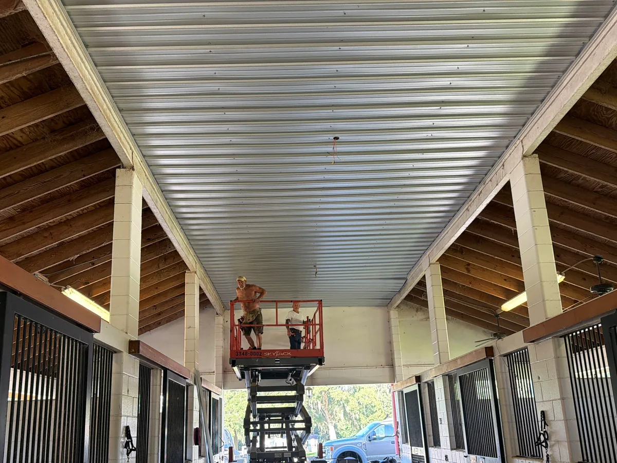 Horse barn aisle with stalls while crew installs a new metal ceiling from a lift