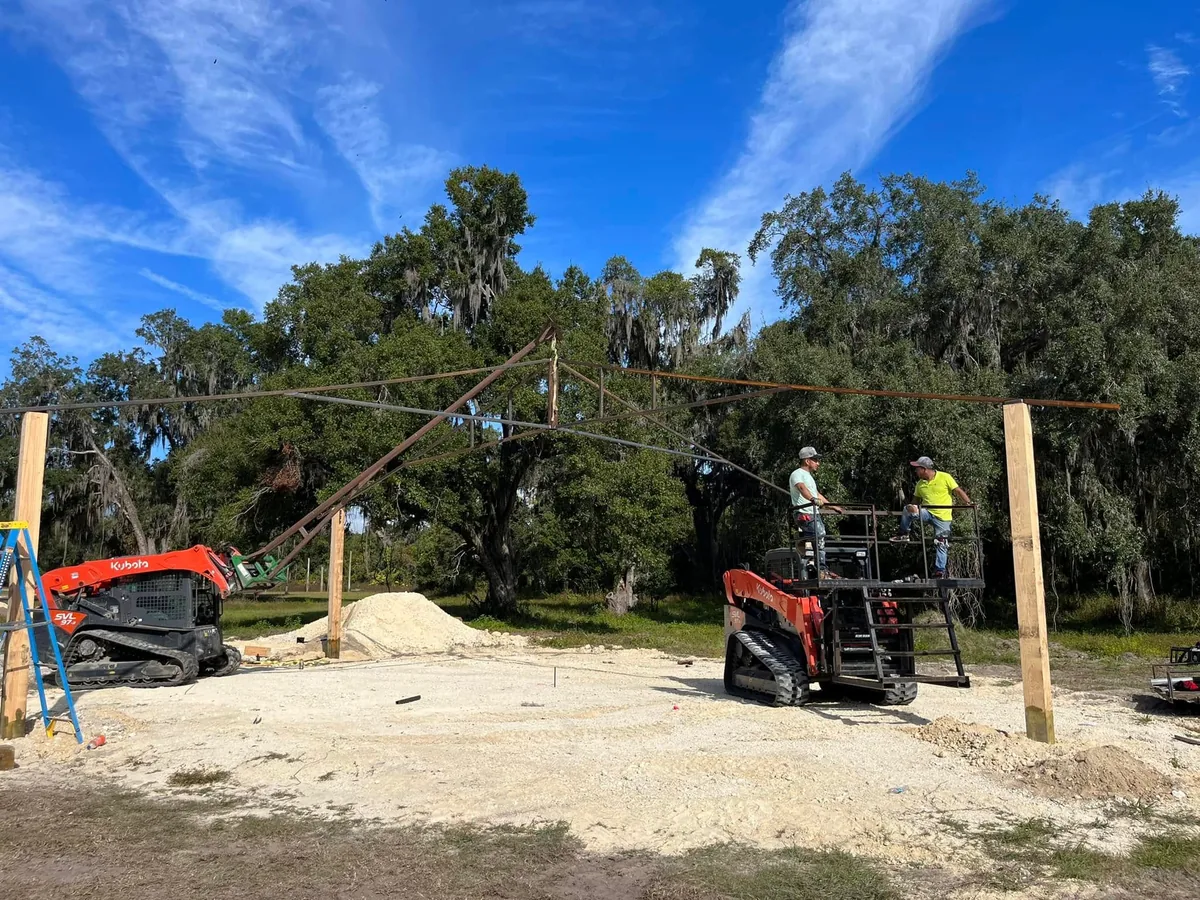 Two track loaders lifting a steel truss into place while crew guides it from a platform
