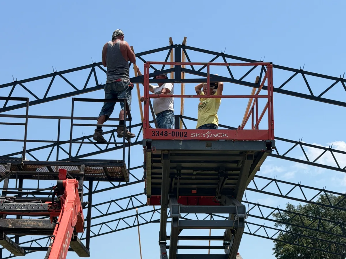 Three crew members on lifts setting black steel trusses on a pole barn frame